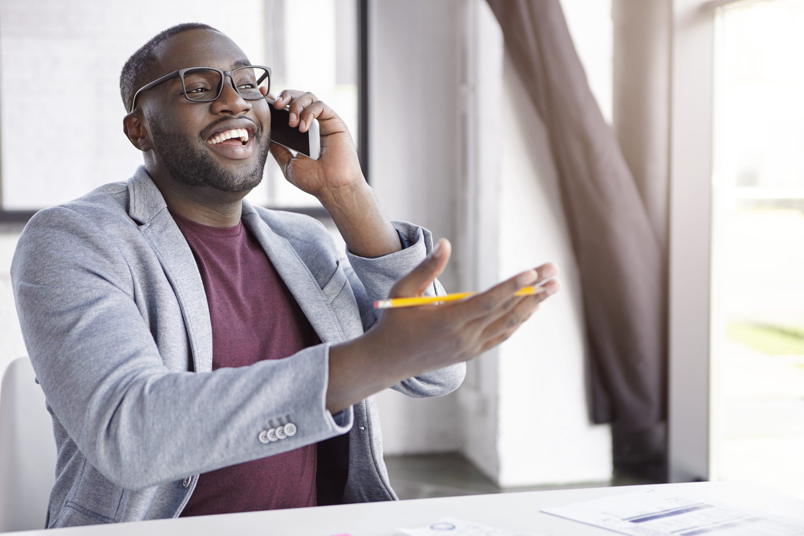 Positive African American male enterpreneur dressed in formal clothes, has conversation over smart phone, gestures actively with hand holds pencil, indicates at something as speaks with friend